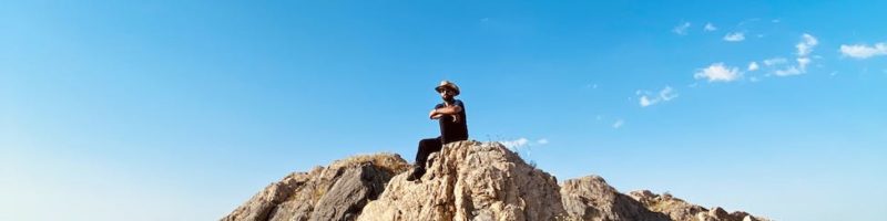man sitting on brown rock formation under blue sky